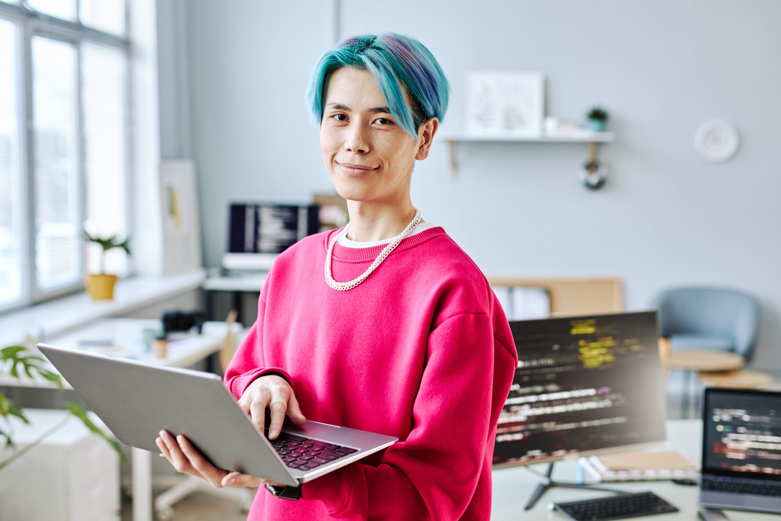 Waist up portrait of Asian young man with colored hair looking at camera in office and holding laptop