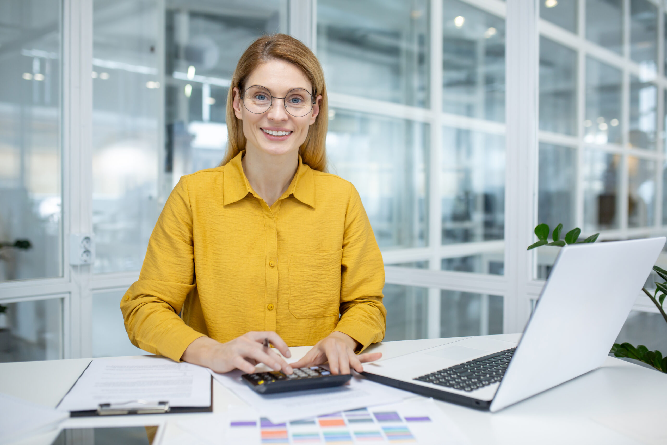Smiling woman in a yellow shirt working at a desk with a laptop, calculator, and documents in a modern office environment.